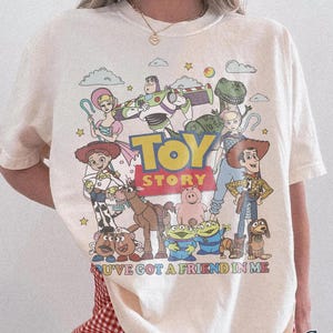 May include: Ivory-colored t-shirt featuring a graphic of Toy Story characters. The design includes Woody, Buzz Lightyear, and other characters from the animated film. The text "TOY STORY" is prominently displayed above the characters, with "YOU'VE GOT A FRIEND IN ME" below.