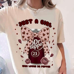 May include: Cream-colored t-shirt with a graphic of a Dr Pepper can adorned with a bow featuring heart patterns. The text reads "JUST A GIRL WHO LOVES DR PEPPER" with the number 23 on the can. Two roses are at the bottom.