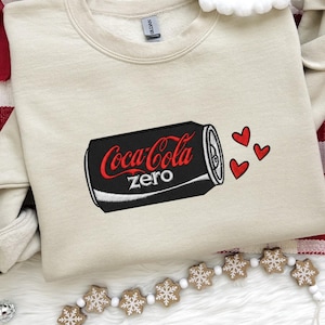 May include: A beige sweatshirt featuring an embroidered Coca-Cola Zero can design with red hearts. The black can has the Coca-Cola Zero logo in red. The sweatshirt is on a white surface with red and white accents.