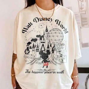 May include: Cream-colored t-shirt featuring a black and white graphic of the Walt Disney World castle, Spaceship Earth, and Mickey Mouse. The text "Walt Disney World" arches across the top, with "The happiest place on earth" below.