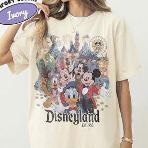 May include: An ivory short-sleeve t-shirt featuring a graphic print of Mickey Mouse, Minnie Mouse, Donald Duck, Goofy, and Pluto in front of a castle and Ferris wheel. The text reads Disneyland Est. 1955. A Comfort Colors Ivory label is visible.