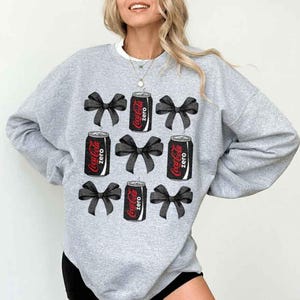 May include: A grey sweatshirt with a design of black bows and Coca-Cola Zero cans. The cans are red and black, with the words "Coca-Cola Zero" printed on them. The bows are arranged around the cans.