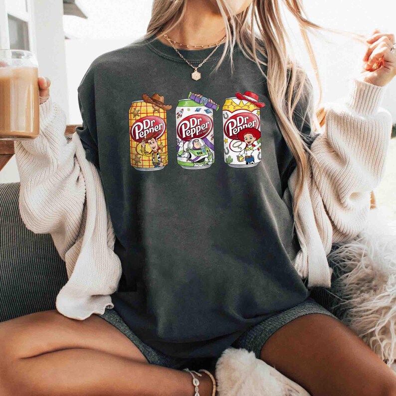 May include: Dark gray sweatshirt featuring three Dr. Pepper soda can designs with cartoon characters. The cans are yellow, green, and white, each with a different character and the Dr. Pepper logo. The sweatshirt is a casual style.