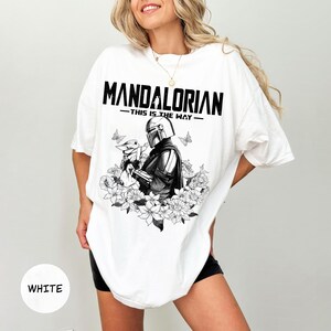 Może przedstawiać: Biała koszulka z czarno-białą grafiką przedstawiającą The Mandalorian i Grogu, otoczonych kwiatami i motylami. Tekst "MANDALORIAN - THIS IS THE WAY" jest wydrukowany na g&oacute;rze czarnymi literami.