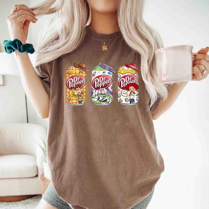 May include: Brown t-shirt featuring three Dr. Pepper soda cans with Toy Story character designs. The cans are yellow, green, and white, each with a different character: Woody, Buzz Lightyear, and Jessie. The shirt has a casual, relaxed fit.