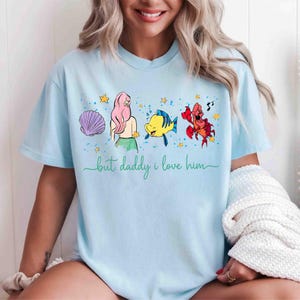 May include: Light blue t-shirt featuring a cartoon design with characters from a popular animated movie. The design includes a seashell, a mermaid, a yellow fish, and a red crab. The text "but daddy i love him" is written in green cursive.
