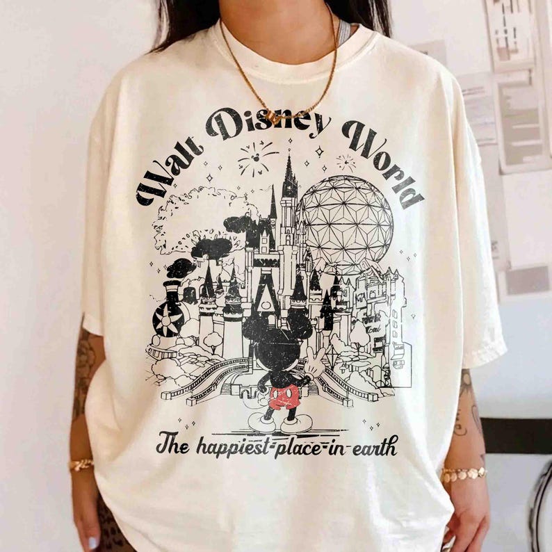 May include: Cream-colored t-shirt featuring a black and white graphic of the Walt Disney World castle, Spaceship Earth, and Mickey Mouse. The text "Walt Disney World" arches across the top, with "The happiest place on earth" below.