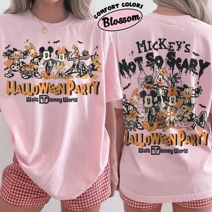 May include: A light pink t-shirt featuring a graphic of Disney characters in skeleton costumes. The front displays the text Halloween Party Walt Disney World. The back shows the same characters with the text Mickey's Not So Scary Halloween Party Walt Disney World. The shirt is labeled Comfort Colors Blossom.