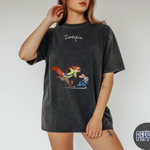 May include: Dark gray t-shirt featuring a cartoon image of a fox and a rabbit from Zootopia. The word "Zootopia" is written above the graphic. The shirt has short sleeves and a relaxed fit.