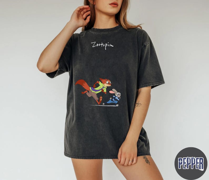 May include: Dark gray t-shirt featuring a cartoon image of a fox and a rabbit from Zootopia. The word "Zootopia" is written above the graphic. The shirt has short sleeves and a relaxed fit.