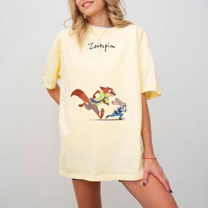 May include: A light yellow t-shirt featuring a cartoon image of Nick Wilde and Judy Hopps from Zootopia. The characters are running, with the word "Zootopia" written above them in black script.