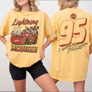 Op de afbeelding: Mosterdgeel T-shirt met een afbeelding van Bliksem McQueen uit de film Cars. Het ontwerp aan de voorkant bevat de tekst "Lightning 95 McQueen" met een afbeelding van de raceauto. De achterkant van het shirt toont een grote "95" en "Lightning McQueen".