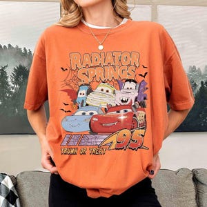 May include: Orange t-shirt featuring a graphic of characters from the movie Cars, including Lightning McQueen, in Halloween costumes. The text "Radiator Springs" and "Trunk or Treat" are also displayed.