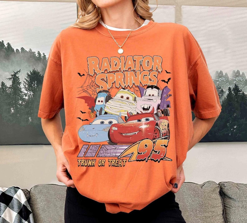 May include: Orange t-shirt featuring a graphic of characters from the movie Cars, including Lightning McQueen, in Halloween costumes. The text "Radiator Springs" and "Trunk or Treat" are also displayed.