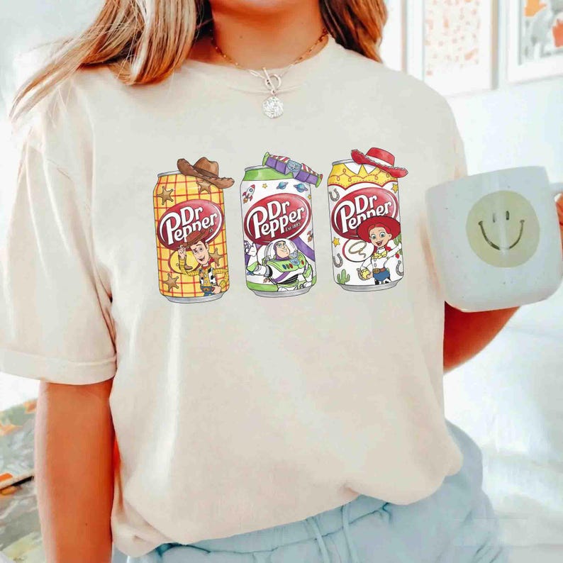 May include: Cream-colored t-shirt featuring three Dr. Pepper soda can designs with Toy Story characters. The cans are decorated with Woody, Buzz Lightyear, and Jessie. A white mug with a smiley face is also visible.