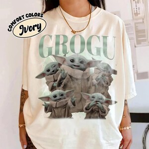 May include: Ivory-colored t-shirt featuring the word "GROGU" in light green, with a graphic of Grogu from The Mandalorian. The graphic shows Grogu in various poses. The shirt has the words "COMFORT COLORS" and "Ivory" in a circular design.