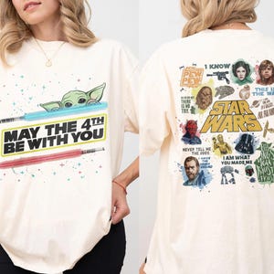 May include: Cream-colored t-shirt featuring a graphic of Baby Yoda with a blue lightsaber and a red lightsaber. The text "MAY THE 4TH BE WITH YOU" is printed below. The back of the shirt has Star Wars characters and quotes.