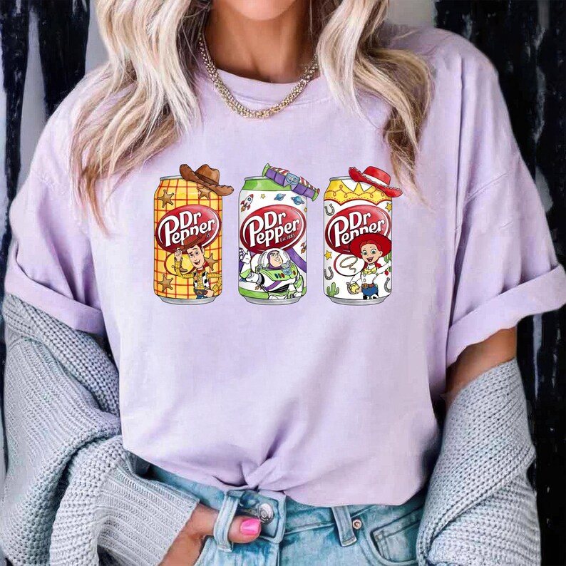 May include: A light purple t-shirt featuring three Dr. Pepper soda cans with cartoon character designs. The cans have cowboy hats and feature characters like Woody and Buzz Lightyear. The Dr. Pepper logo is prominently displayed.