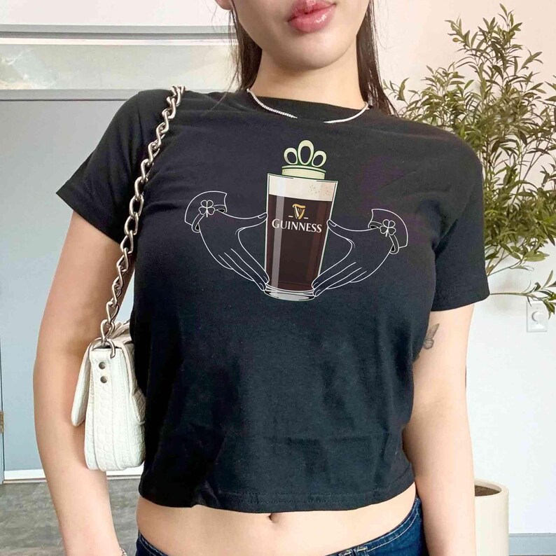 May include: Black crop top featuring a graphic of a pint of Guinness beer held by two hands. The Guinness logo is visible on the glass. The top has short sleeves and a round neckline. A white chain strap bag is visible on the left side.