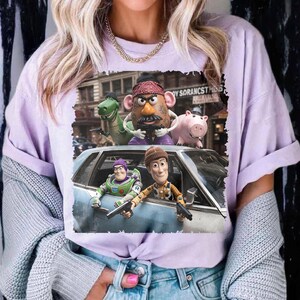 May include: A light purple t-shirt featuring a graphic print of Toy Story characters including Woody, Buzz Lightyear, Mr. Potato Head, Rex, and Hamm. The characters are depicted in a city scene with a car, and the text reads "TOY SORANCST".