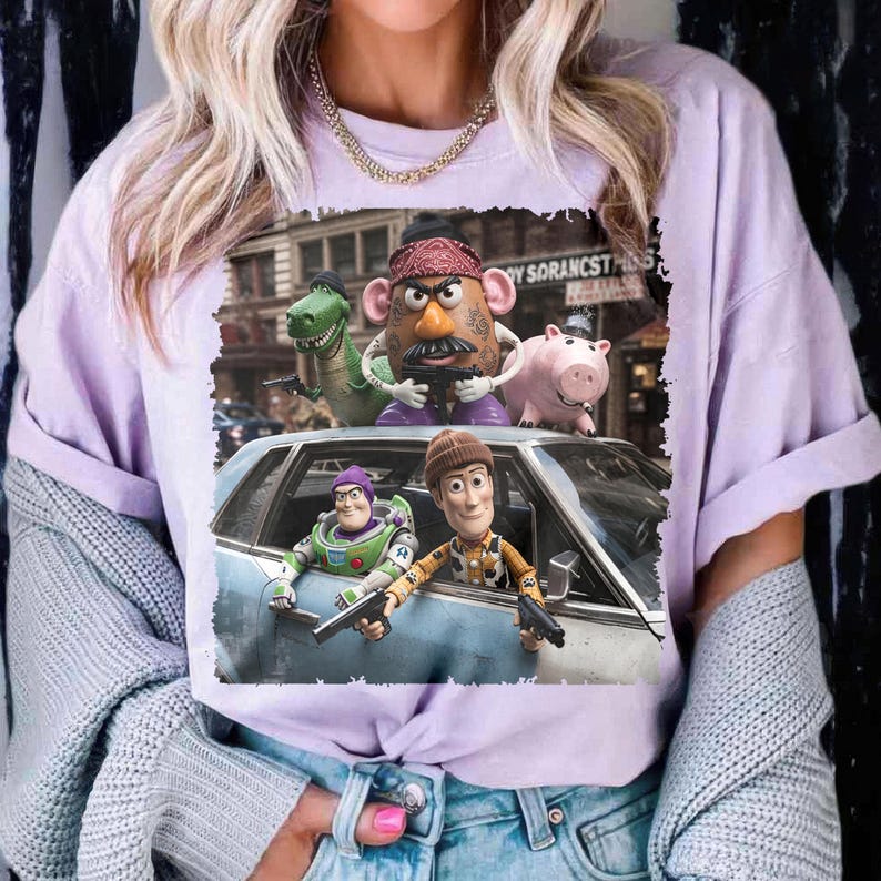 May include: A light purple t-shirt featuring a graphic print of Toy Story characters including Woody, Buzz Lightyear, Mr. Potato Head, Rex, and Hamm. The characters are depicted in a city scene with a car, and the text reads "TOY SORANCST".