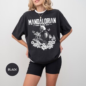 Może przedstawiać: Czarny t-shirt z napisem "MANDALORIAN" i frazą "THIS IS THE WAY". Koszulka przedstawia ilustrację postaci z serialu, otoczoną białymi kwiatami i motylami.