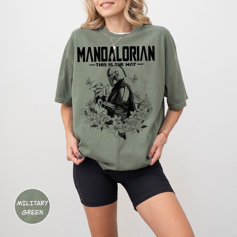 Może przedstawiać: Wojskowa zielona koszulka z napisem "MANDALORIAN - THIS IS THE WAY" nad czarno-białą grafiką Mandalorianina i Grogu otoczonych kwiatami i motylami. Koszulka ma luźny kr&oacute;j.