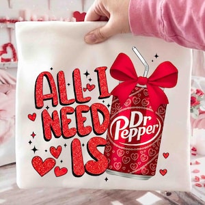 May include: A white t-shirt with the text "ALL I NEED IS" in red glitter letters, accompanied by red hearts and a Dr. Pepper can with a red bow. The Dr. Pepper can has a straw and is decorated with hearts.
