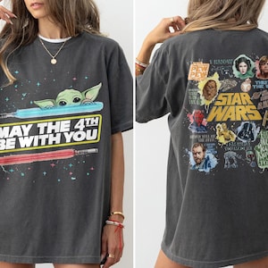 May include: Dark gray t-shirt featuring a graphic of Baby Yoda, lightsabers, and the text "MAY THE 4TH BE WITH YOU." The back of the shirt displays various Star Wars characters and the words "STAR WARS."