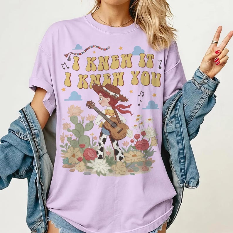 May include: A light purple t-shirt featuring a graphic print of a cowgirl with a guitar standing among cacti and flowers. The text "I KNEW IT I KNEW YOU" is printed in a yellow retro font above the character, accompanied by small stars and clouds.