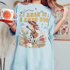 May include: A light blue t-shirt featuring a graphic of a cowgirl playing a guitar in a field of cacti and flowers. The text "I KNEW IT I KNEW YOU" is printed above the character in a retro font, accompanied by musical notes and clouds.