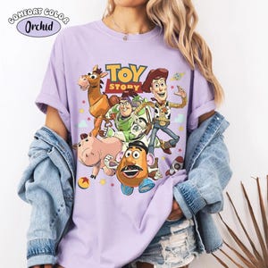 May include: A light purple t-shirt featuring a graphic print of Toy Story characters including Woody, Buzz Lightyear, Bullseye, Hamm, and Mr. Potato Head. The text "TOY STORY" is printed in yellow and red above the characters, with small stars and a ball.