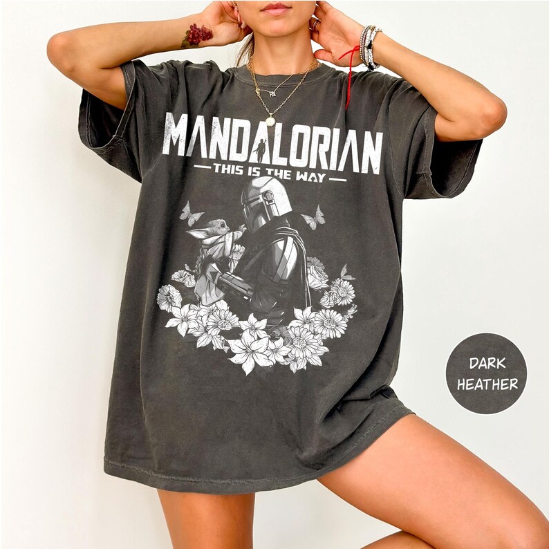 Może przedstawiać: Ciemnoszara koszulka z białym napisem "MANDALORIAN - THIS IS THE WAY". Grafika przedstawia Mandalorianina i Grogu otoczonych kwiatami i motylami. Koszulka ma luźny kr&oacute;j.