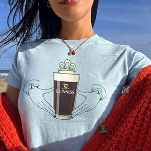 May include: Light blue t-shirt featuring a Guinness pint design. The graphic includes a pint of dark beer held by two hands with a crown above the glass. The word "Guinness" is printed on the glass. The person is wearing a red sweater.