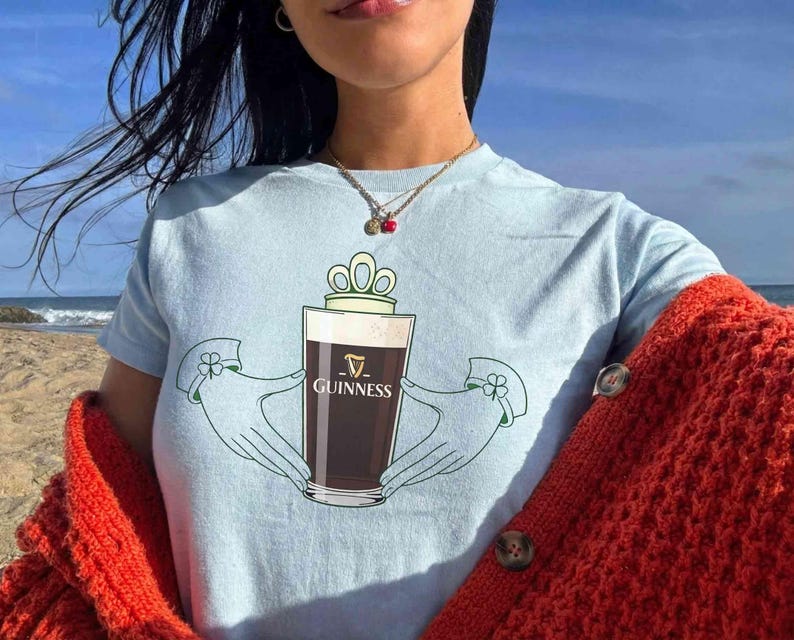 May include: Light blue t-shirt featuring a Guinness pint design. The graphic includes a pint of dark beer held by two hands with a crown above the glass. The word "Guinness" is printed on the glass. The person is wearing a red sweater.