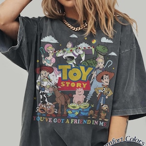以下が含まれることがあります： アニメ映画「トイ・ストーリー」のキャラクターが描かれた、ダークグレーのTシャツ。ウッディ、バズ・ライトイヤーなどのキャラクターと、「TOY STORY」と「YOU'VE GOT A FRIEND IN ME」の文字がデザインされています。
