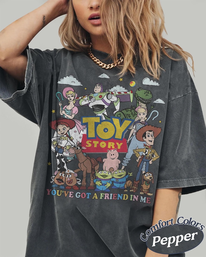 以下が含まれることがあります： アニメ映画「トイ・ストーリー」のキャラクターが描かれた、ダークグレーのTシャツ。ウッディ、バズ・ライトイヤーなどのキャラクターと、「TOY STORY」と「YOU'VE GOT A FRIEND IN ME」の文字がデザインされています。