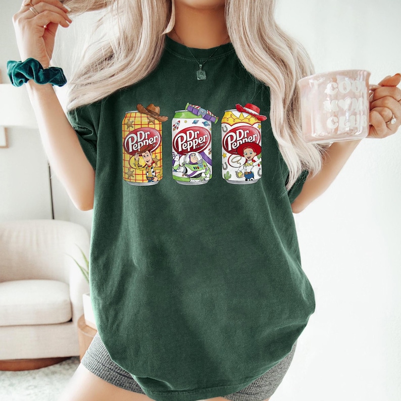 May include: Dark green t-shirt featuring three Dr. Pepper soda cans with cartoon character designs. The cans have cowboy hats and character illustrations. The shirt is a comfortable, casual style.