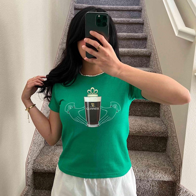 May include: Green t-shirt featuring a Guinness pint design. The graphic includes a pint of dark beer held by two hands, with a crown above the glass. The word "Guinness" is printed on the glass. The shirt is short-sleeved.