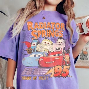 May include: A purple t-shirt featuring a graphic of the Radiator Springs characters from the movie Cars, dressed in Halloween costumes. The text "Radiator Springs" and "Trunk or Treat" are printed in orange and white.