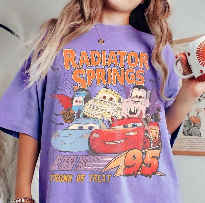 May include: A purple t-shirt featuring a graphic of the Radiator Springs characters from the movie Cars, dressed in Halloween costumes. The text "Radiator Springs" and "Trunk or Treat" are printed in orange and white.