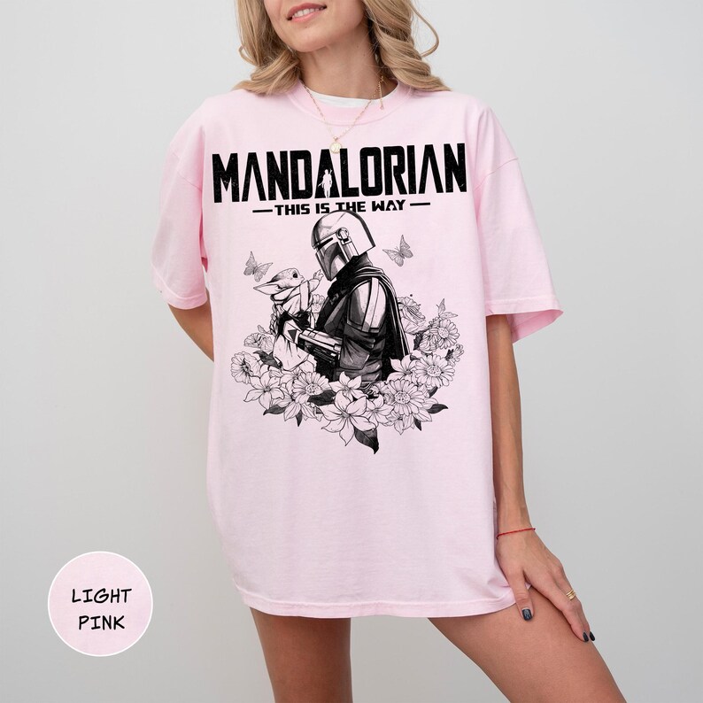 Może przedstawiać: Jasnor&oacute;żowa koszulka z czarno-białą grafiką przedstawiającą The Mandalorian i Grogu otoczonych kwiatami i motylami. Tekst "MANDALORIAN - THIS IS THE WAY" jest wydrukowany nad grafiką.