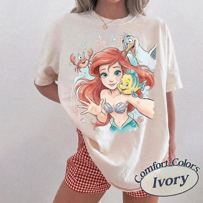 May include: An ivory-colored short-sleeve t-shirt featuring a graphic print of a red-haired mermaid character, a small yellow and blue fish, a red crab, and a white seagull. The design includes light blue bubbles and coral accents.