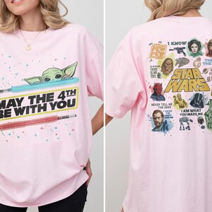 May include: A light pink Star Wars t-shirt. The front features Baby Yoda, a blue lightsaber, a red lightsaber, and the text "MAY THE 4TH BE WITH YOU." The back has various Star Wars characters and quotes.