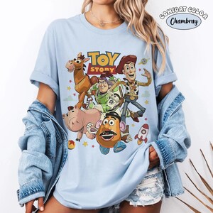 May include: A light blue short-sleeve t-shirt featuring a graphic print of Toy Story characters including Woody, Buzz Lightyear, Bullseye, Hamm, and Mr. Potato Head. The text Toy Story is printed above the characters with small stars and a rocket.