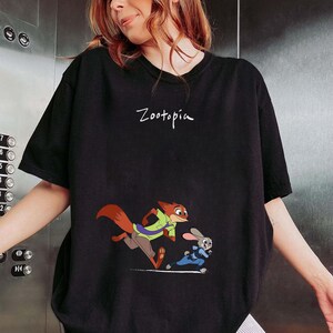 May include: Black t-shirt featuring the word "Zootopia" in white script. The shirt displays a cartoon image of a fox and a rabbit running. The fox is wearing a green shirt and brown pants, and the rabbit is wearing a blue uniform.