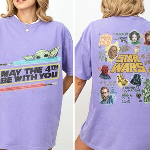 May include: A lilac-colored t-shirt featuring a Star Wars theme. The front displays Baby Yoda with lightsabers and the text "MAY THE 4TH BE WITH YOU." The back showcases various characters and phrases from the Star Wars saga.