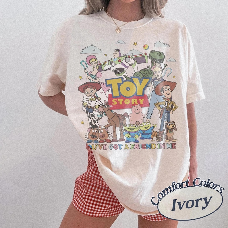 May include: Ivory-colored t-shirt featuring a graphic of Toy Story characters. The design includes Woody, Buzz Lightyear, and other characters from the animated film. The text "TOY STORY" is prominently displayed above the characters, with "YOU'VE GOT A FRIEND IN ME" below.