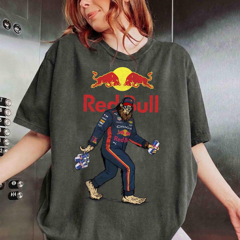 Pu&ograve; includere: T-shirt grigio scuro con un personaggio simile a Bigfoot in tuta da corsa Red Bull, con lattine Red Bull. Il logo e il testo Red Bull sono sopra il personaggio. La maglietta ha un aspetto vintage.