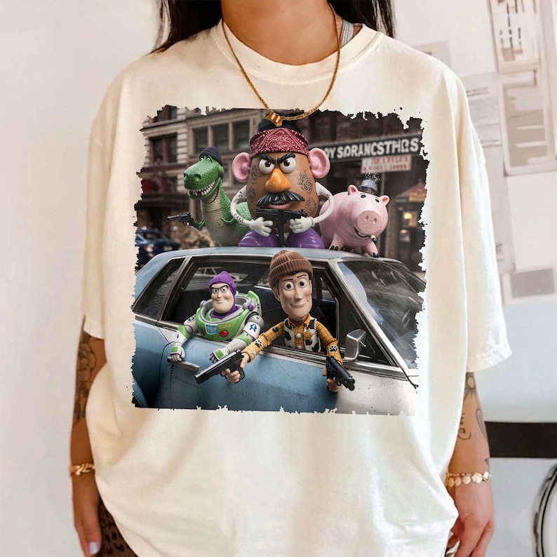 May include: A cream-colored t-shirt featuring a graphic print of Toy Story characters including Woody, Buzz Lightyear, Mr. Potato Head, Rex, and Hamm. The characters are depicted in a scene with a blue car and a city background, with some holding toy guns.